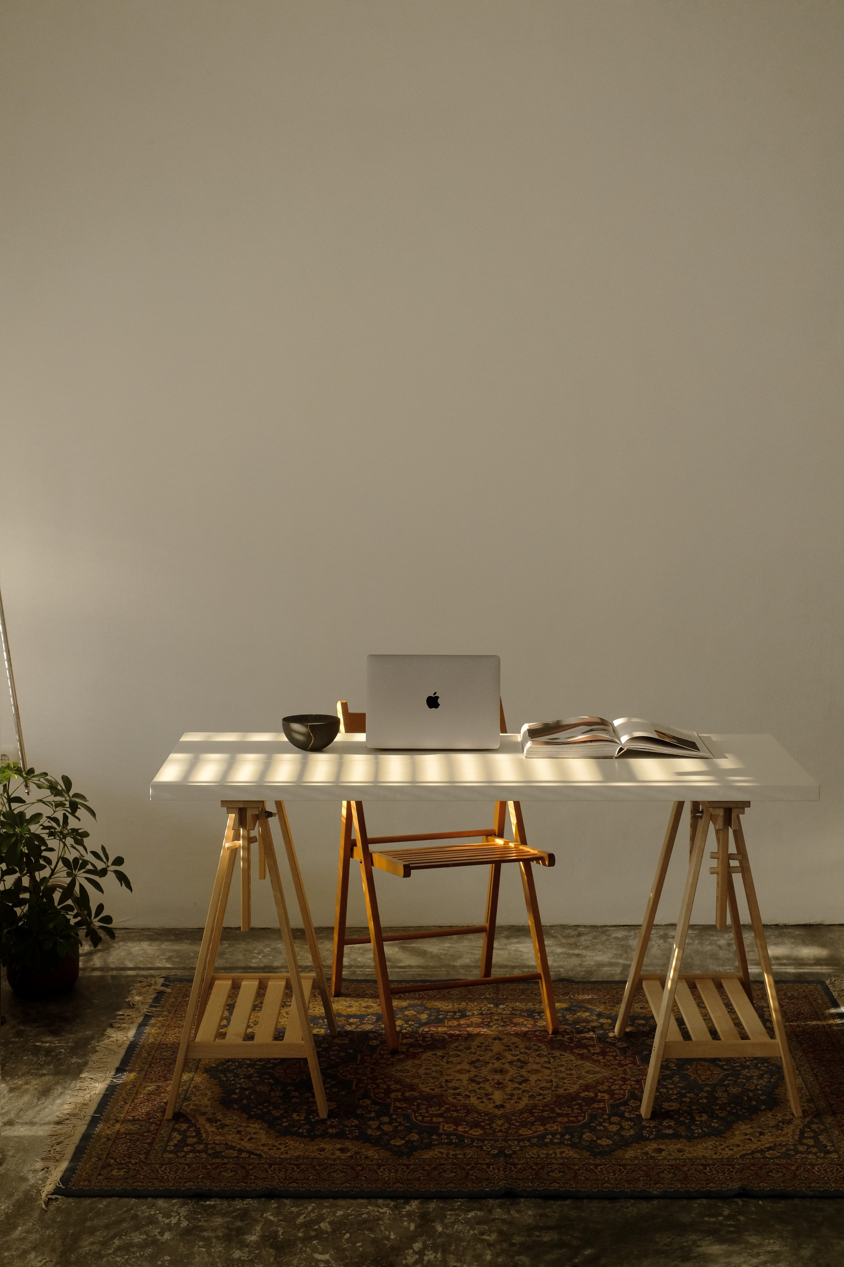 A sunlit studio desk with a laptop, an open book and a wooden chair